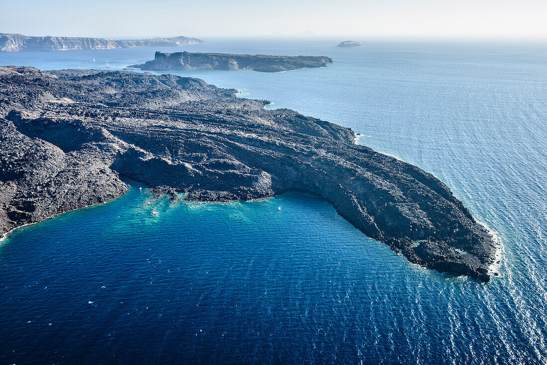 Aerial view of a headland on a island in the Aegean sea, volcanic rock formations, black ridges and small quiet inlets and islands. Boats in the distance.