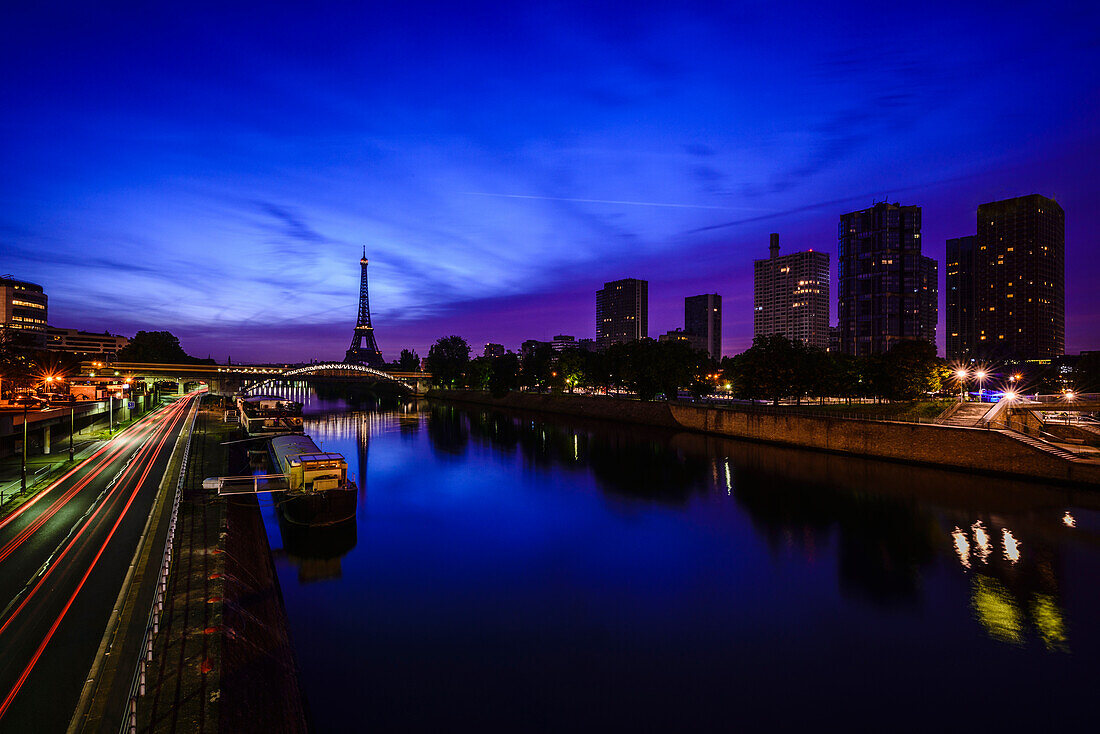 A view along the water of the River Seine at night, tall buildings on the river bank, the Eiffel tower in the distance.