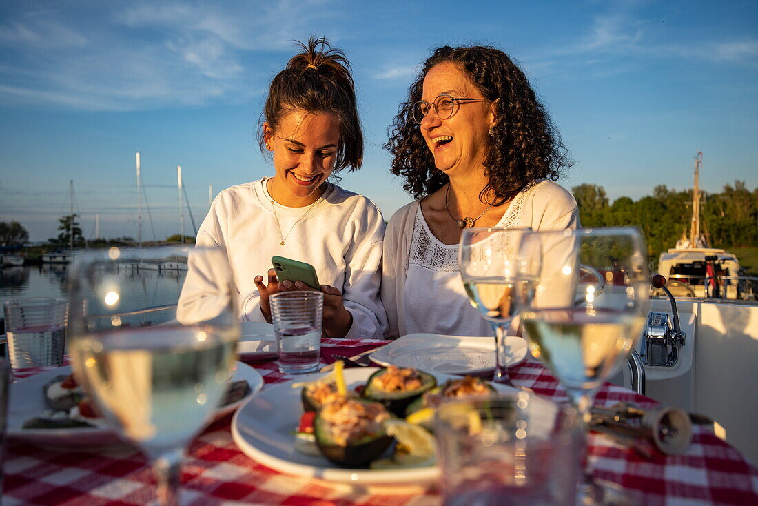 Junge erwachsene Tochter und Mutter genießen Abendessen an Deck von einem Le Boat Elegance Hausboot, See De Fluezen, Friesland, Niederlande, Europa