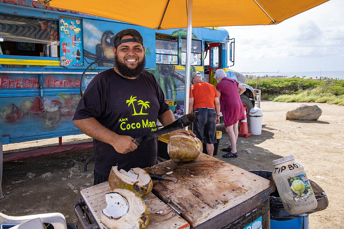 Freundlicher Mann öffnet frische Kokosnüsse an einer Snackbar, alter Schulbus am California Lighthouse, Noord, Aruba, Niederländische Karibik, Karibik