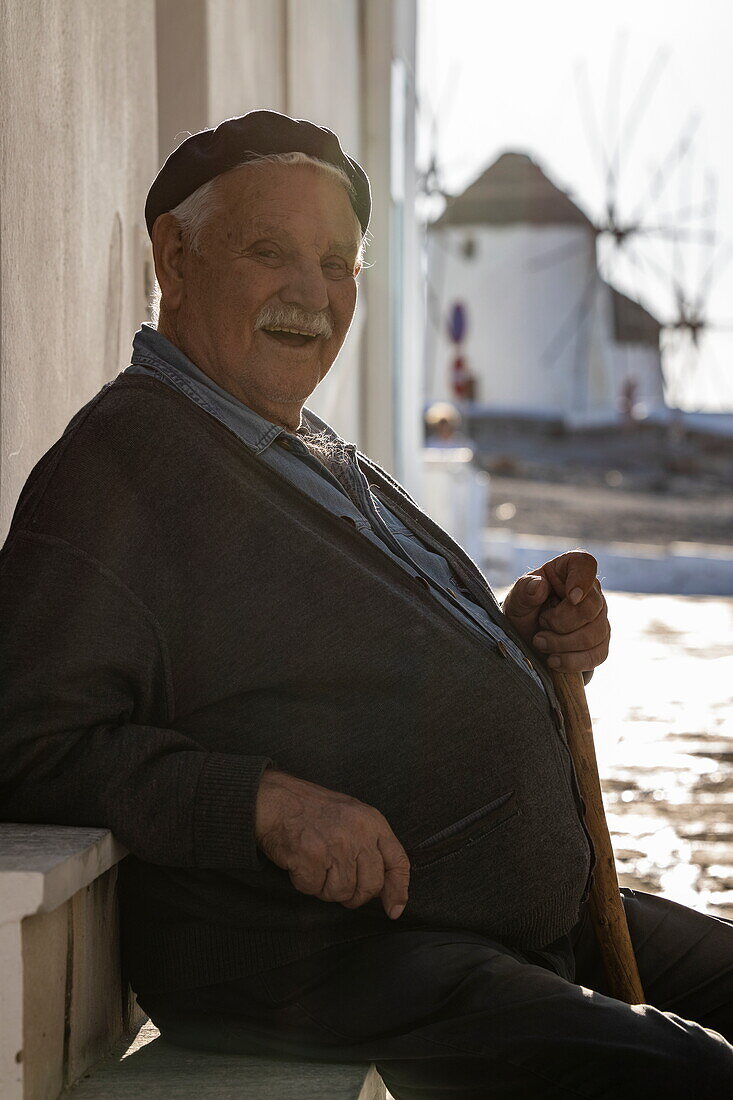 Würdevoller älterer Mann sitzt auf Stufe in einer Gasse mit den berühmten Windmühlen von Mykonos dahinter, Mykonos, Südliche Ägäis, Griechenland, Europa