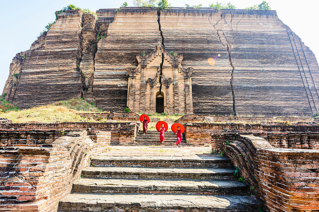Three monks with orange umbrellas outside a large rock temple, the entrance carved into the rock face at Saigang.