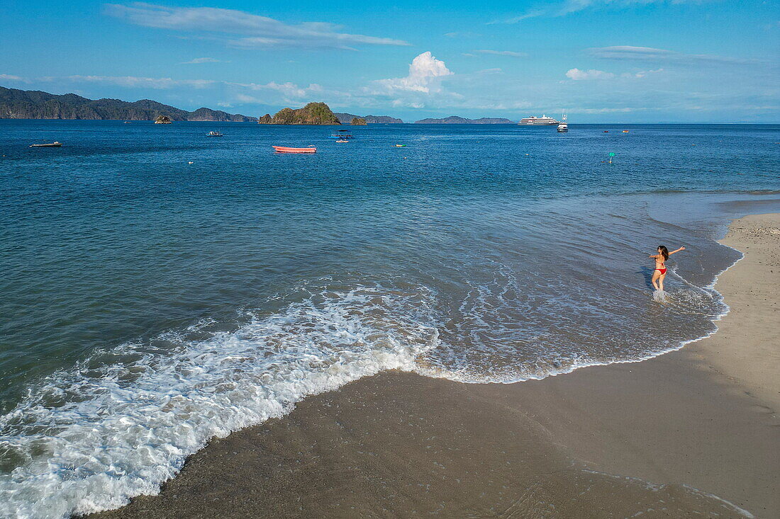 Luftaufnahme einer Frau die am Strand spaziert mit Expeditionskreuzfahrtschiff World Voyager (nicko cruises) in der Ferne, Isla Tortuga, Puntarenas, Costa Rica, Mittelamerika