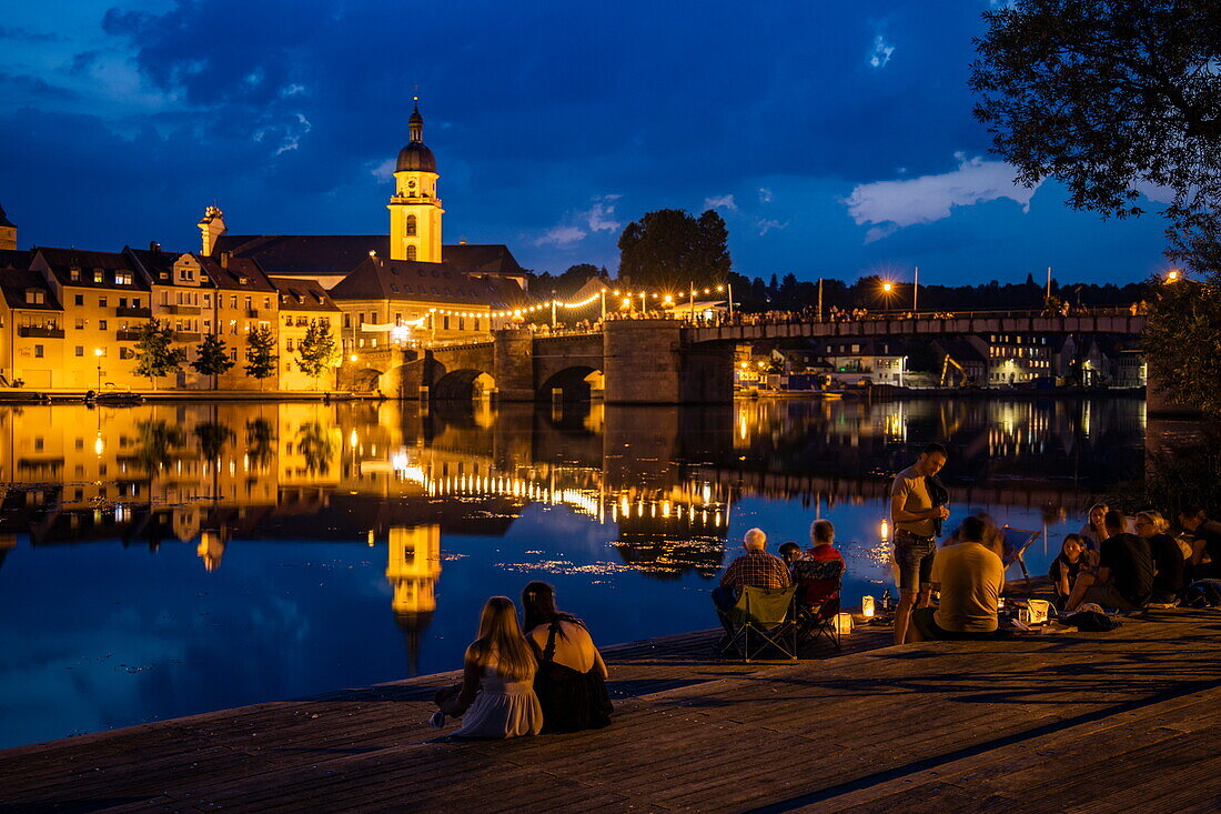 Menschen entspannen am Flussufer mit Spiegelung von Kirchen und Brücke im Main bei Nacht, Kitzingen, Franken, Bayern, Deutschland, Europa