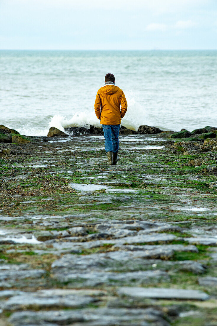 Frau in orangefarbener Jacke und blauer Hose, die auf das Meer zugeht.