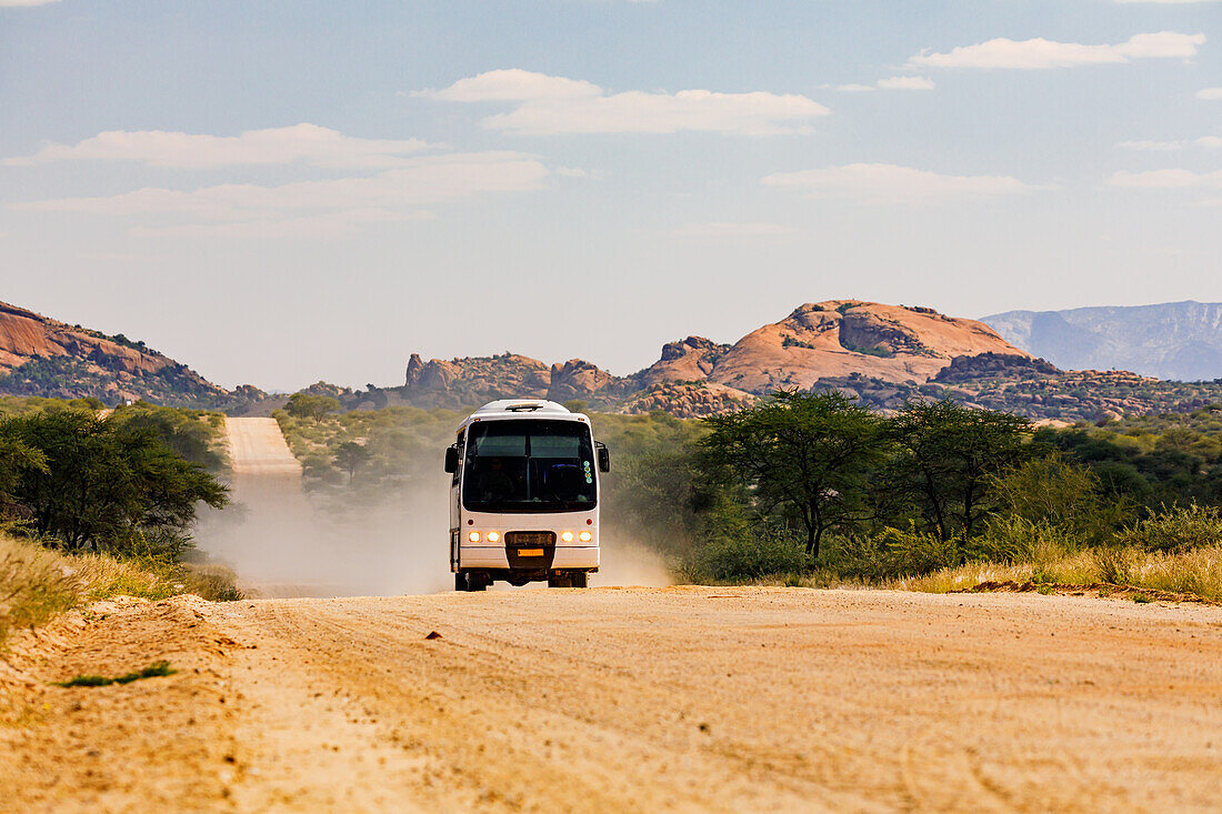 Ein moderner Bus für Touristen fährt auf einer staubigen Schotterpiste durch die Felsen im Erongo Gebirge, Namibia, Afrika