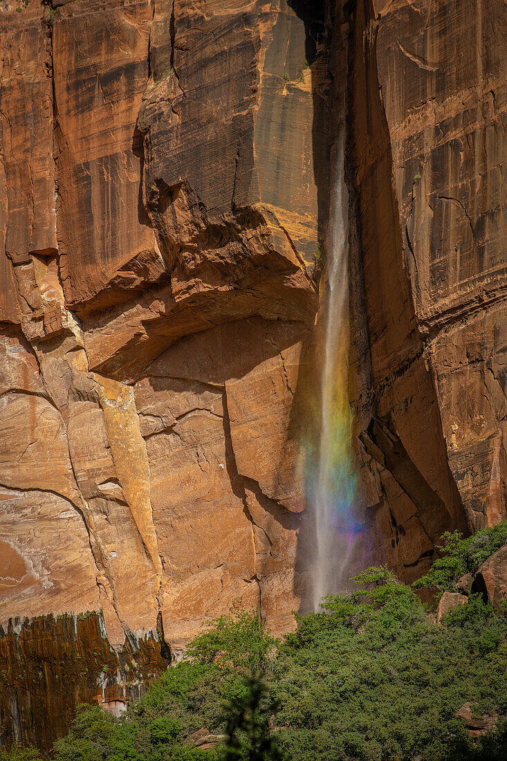 Small waterfall in front of a red rock face is illuminated by the sun and shines in rainbow colors.