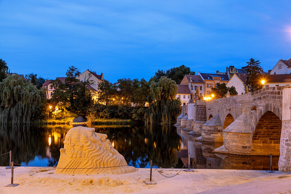 Steinbrücke Kamenný Most über den Fluss Otava im Abendlicht mit Sandskulptur am Ufer in Písek in Südböhmen in Tschechien