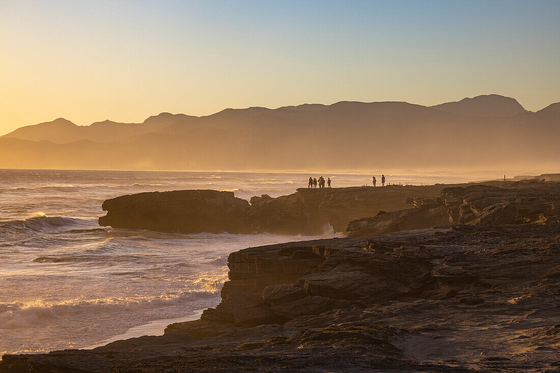 People with fat tire bicycles stand on rocks as waves break onto the coast and beach at sunset in Walker Bay Nature Reserve, Gansbaai De Kelders, Western Cape, South Africa