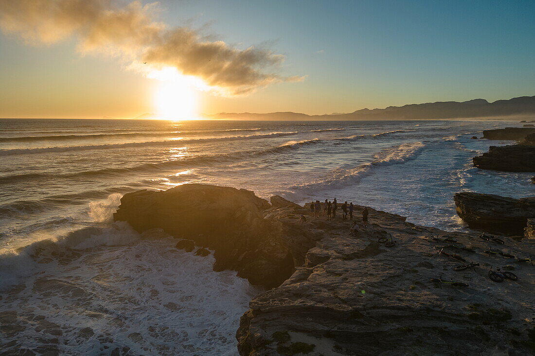 Aerial view of people standing on a rocky outcrop with fat tire bicycles as waves break onto the coast and beach at sunset in Walker Bay Nature Reserve, Gansbaai De Kelders, Western Cape, South Africa