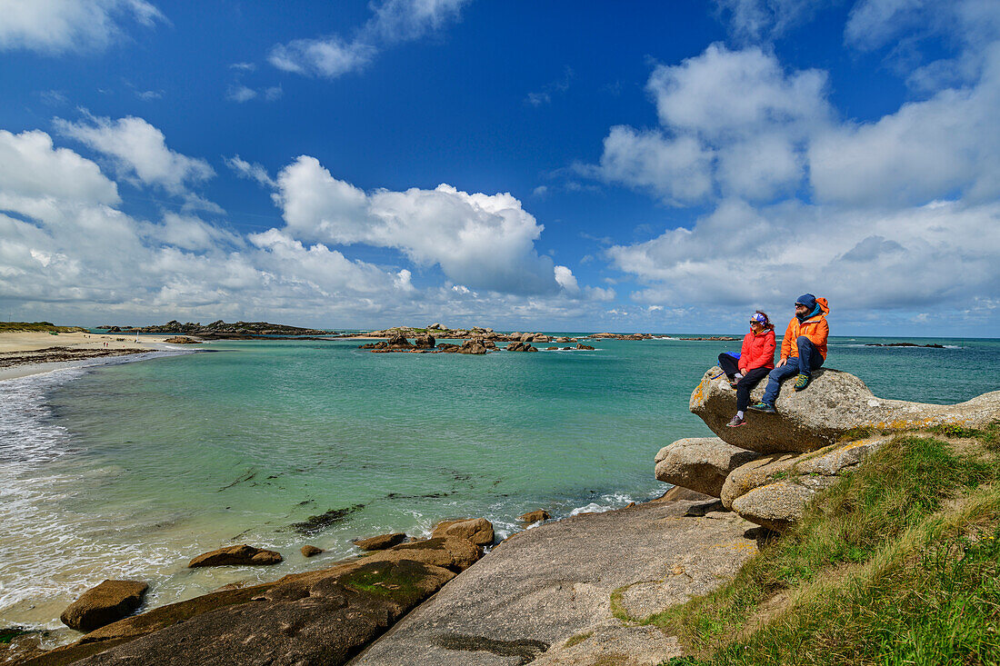 Man and woman hiking sitting on rocks and looking at sandy beach, Trégastel, Côte de Granit Rose, Brittany, France