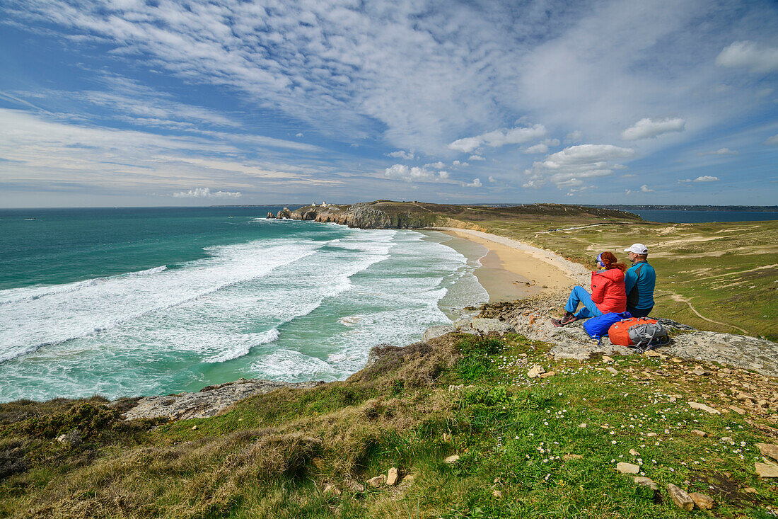Man and woman hiking sitting on rocks and looking at Plage de Pen Hat beach, Camaret-sur-Mer, GR 34, Zöllnerweg, Sentier Côtier, Crozon peninsula, Atlantic coast, Brittany, France