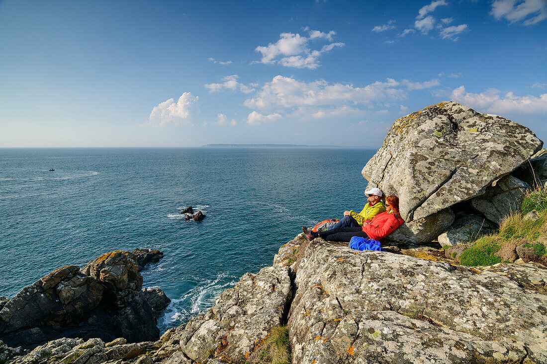 Mann und Frau beim Wandern sitzen auf Felsen und blicken auf die Küste, Cap-Sizun, GR 34, Zöllnerweg, Bretagne, Frankreich