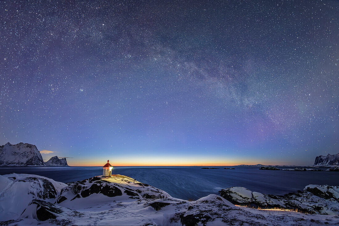 Beleuchteter Leuchtturm steht auf Landzunge mit verschneiten Bergen und Sternhimmel mit Milchstraße, Senja, Troms og Finnmark, Norwegen