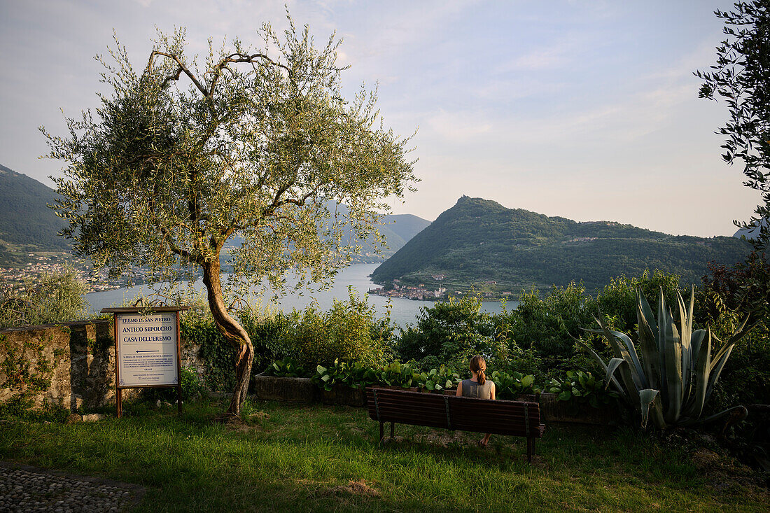 Frau sitzt auf Bank und blickt zur Insel "Monte Isola" im Iseosee (Lago d'Iseo, auch Sebino), Vesto, Brescia und Bergamo, Oberitalienische Seen, Lombardei, Italien, Europa