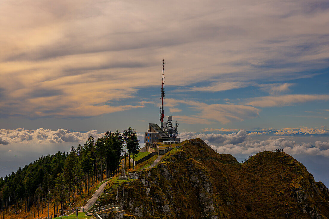 Wetter- und Kommunikationsstation mit Bäumen auf dem Berggipfel und Wolkengebilde an einem sonnigen Tag in Monte Generoso, Tessin, Schweiz.