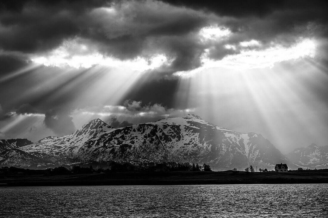Sonnenstrahlen scheinen durch Wolkenloch auf Berge, Fjord und einzelnes Haus, Skreda, Lofoten, Nordland, Norwegen