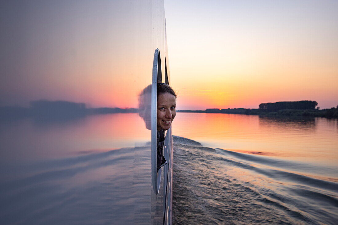 Lächelnde Frau schaut durch Fensteröffnung an Bord von Flusskreuzfahrtschiff Maxima (nicko cruises) auf der Donau bei Sonnenuntergang, in der Nähe von Golubac, Rumänien, Europa