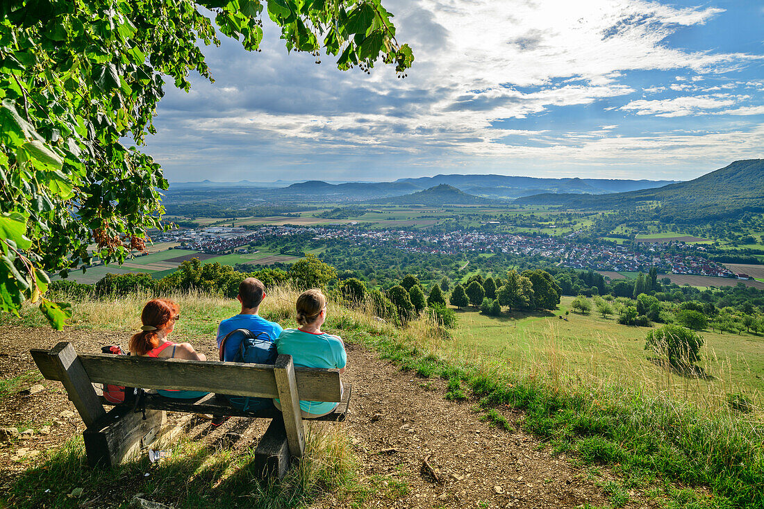 Three people hiking sit on a bench and look towards Bissingen, from the Hörnle, Teck, Swabian Alb, Baden-Württemberg, Germany