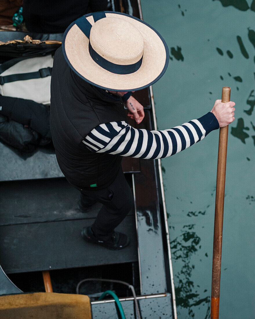 Ein Gondoliere fährt mit dem traditionellen Canotier-Strohhut und einem gestreiften Pullover durch die Kanäle von Venedig in Italien