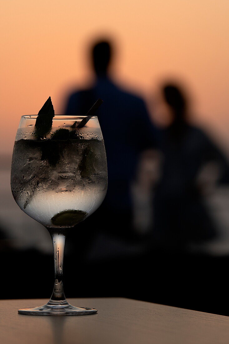  Detail of a gin and tonic cocktail served aboard the expedition cruise ship SH Diana (Swan Hellenic) with a couple in silhouette at sunset, at sea, near Yemen, Middle East 