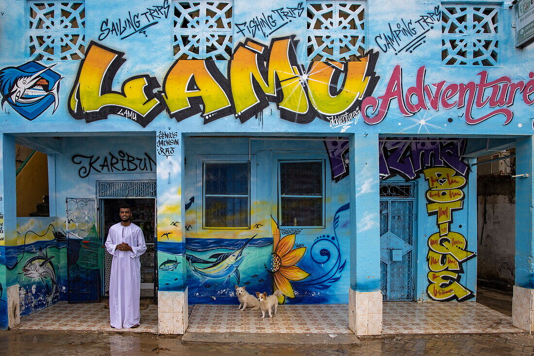 Mann in traditionellem Umhang und Hunde vor dem Geschäft und Tourbüro von Lamu Adventures, Lamu, Insel Lamu, Kenia, Afrika