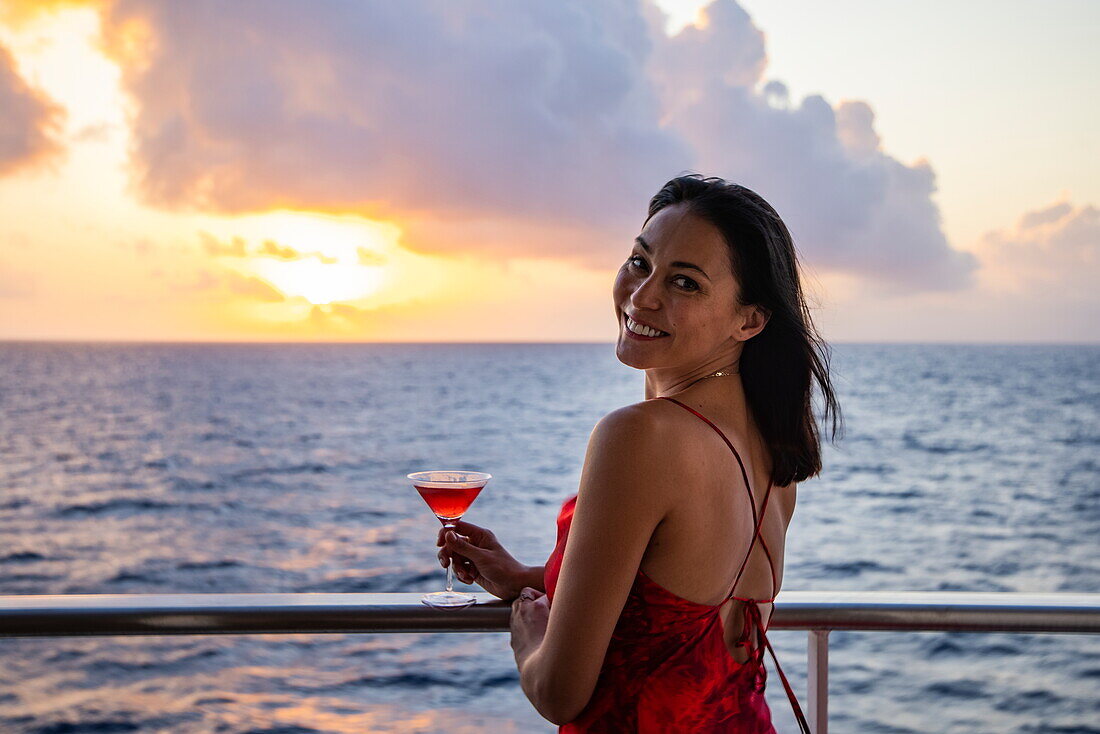 Lächelnde Frau mit Cocktailglas an der Reling des Expeditionskreuzfahrtschiffes SH Diana (Swan Hellenic) bei Sonnenuntergang, in der Nähe der Seychellen, Indischer Ozean