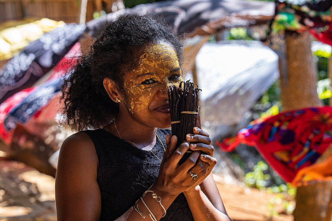  Happy woman with Masonjoany face paint as sunscreen smells vanilla beans for sale at local market, Nosy Komba, Diana, Madagascar, Indian Ocean 