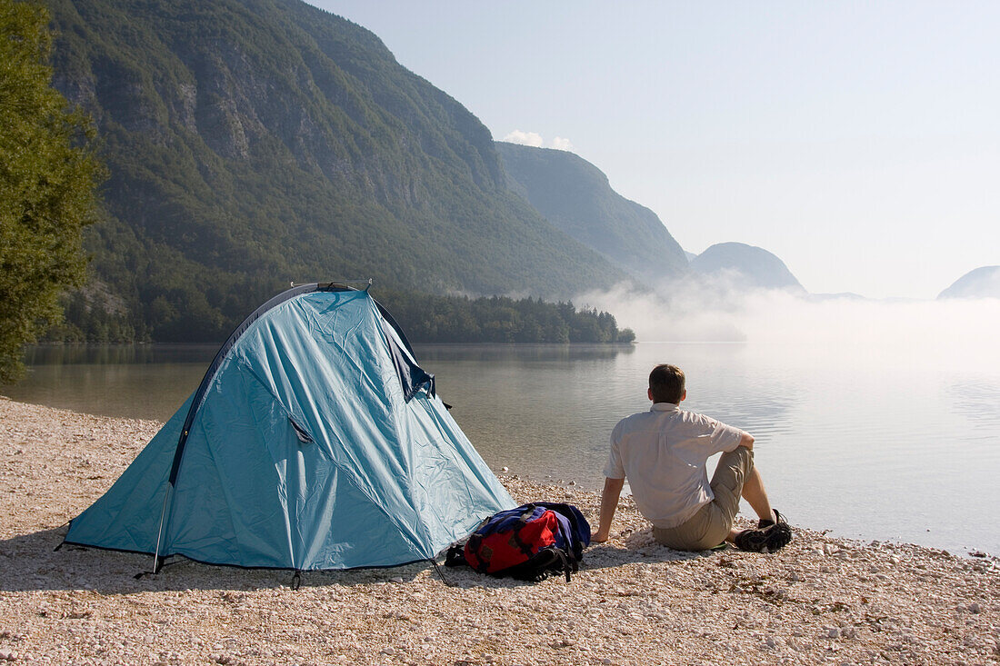 Mann sitzt neben seinem Zelt an einem See im Triglav Nationalpark, Ukanc, Slowenien