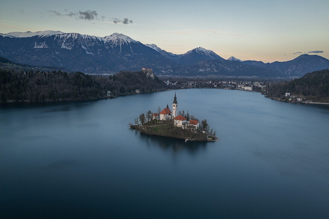 Vogelperspektive der Marienkirche im Bleder See und der verschneiten Berge kurz nach Sonnenuntergang in Bled, Slowenien, Europa.