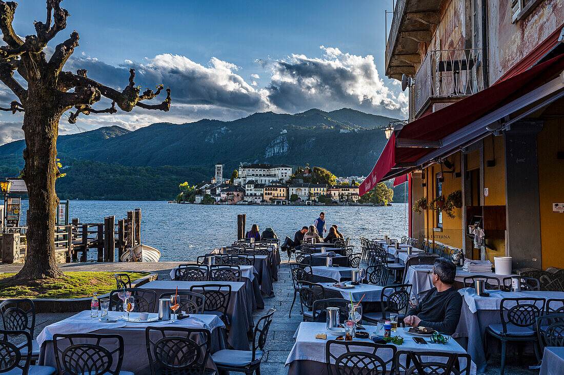  View of Isola San Giulio from the port of Orta San Giulio, Piazza Motta, tables and chairs in front of restaurant, Hotel Leon d&#39;Oro left side, Orta San Giulio, Lake Orta is a northern Italian lake in the northern Italian, Lago d&#39;Orta, or Cusio, region of Piedmont, Italy, Europe 