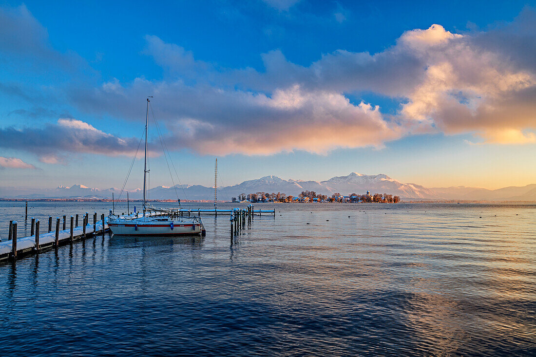 Verschneiter Steg mit Segelboot am Chiemsee mit Fraueninsel und Chiemgauer Alpen im Hintergrund, Chiemsee, Chiemgauer Alpen, Oberbayern, Bayern, Deutschland