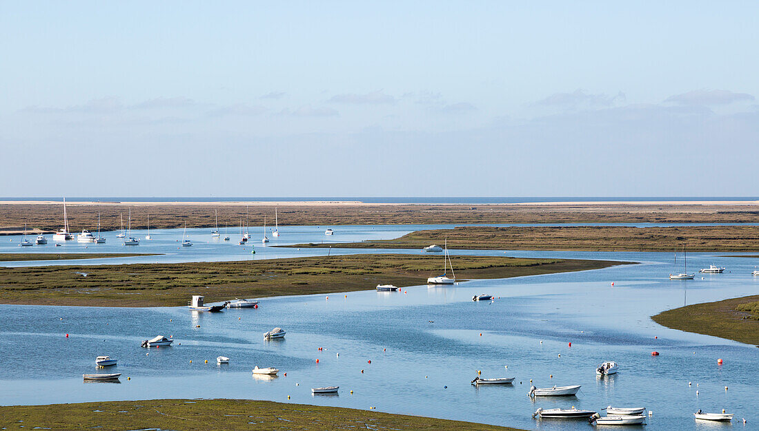 Coastal landscape of salt marsh with boats moored in meandering river drainage channels along the coastline off Faro, Algarve, Portugal