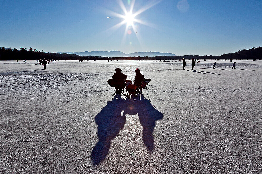 Pärchen sitzt auf Klappstühlen auf zugefrorenem See neben Fußgänger und Eisläufer, Südtirol, Trentino, Italien