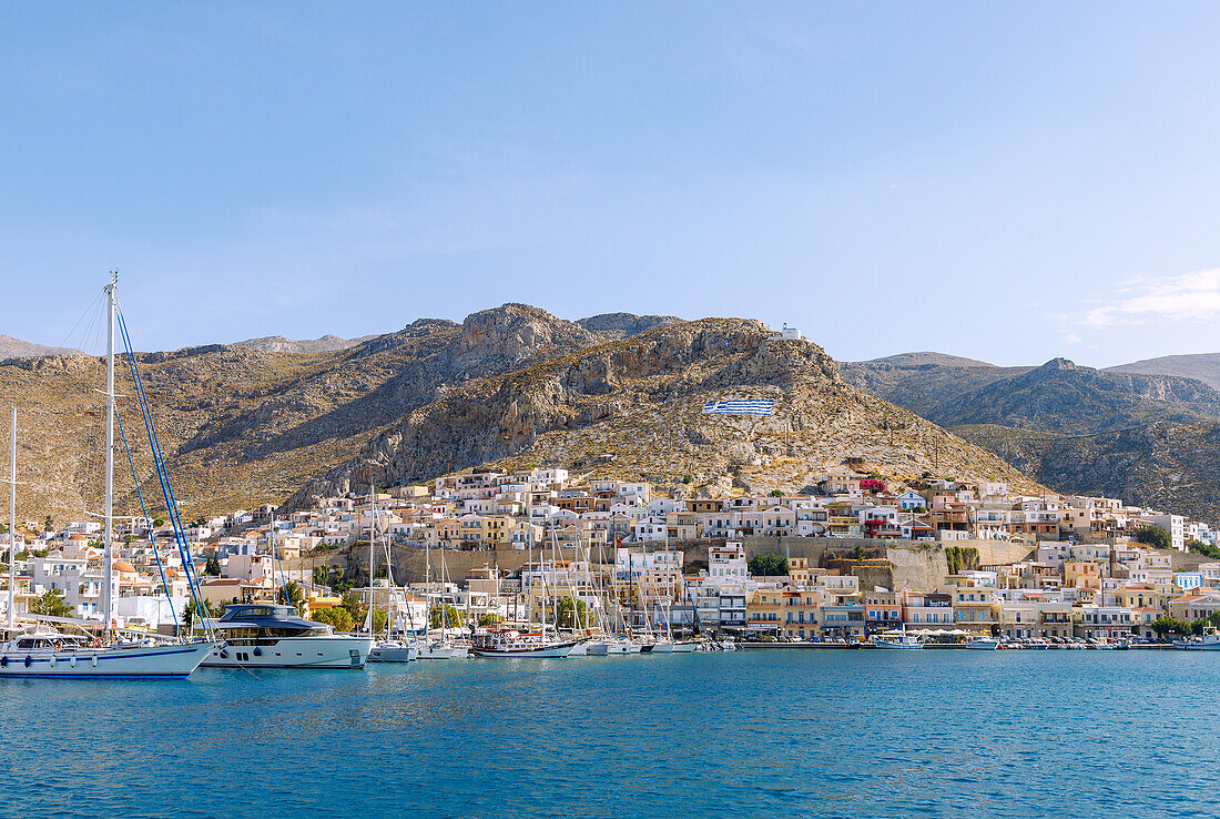 Inselhauptstadt Póthia, weiße Kapelle, griechische Flagge, pastellfarbene Häuser und Boote im Hafen auf der Insel Kalymnos (Kalimnos) in Griechenland