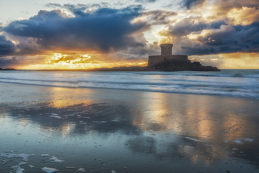  La Rocco Tower stands on the sandy beach at sunset. The sky is reflected in the water. 