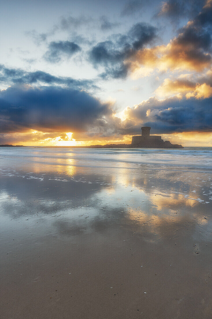  La Rocco Tower stands on the sandy beach at sunset. The sky is reflected in the water. 