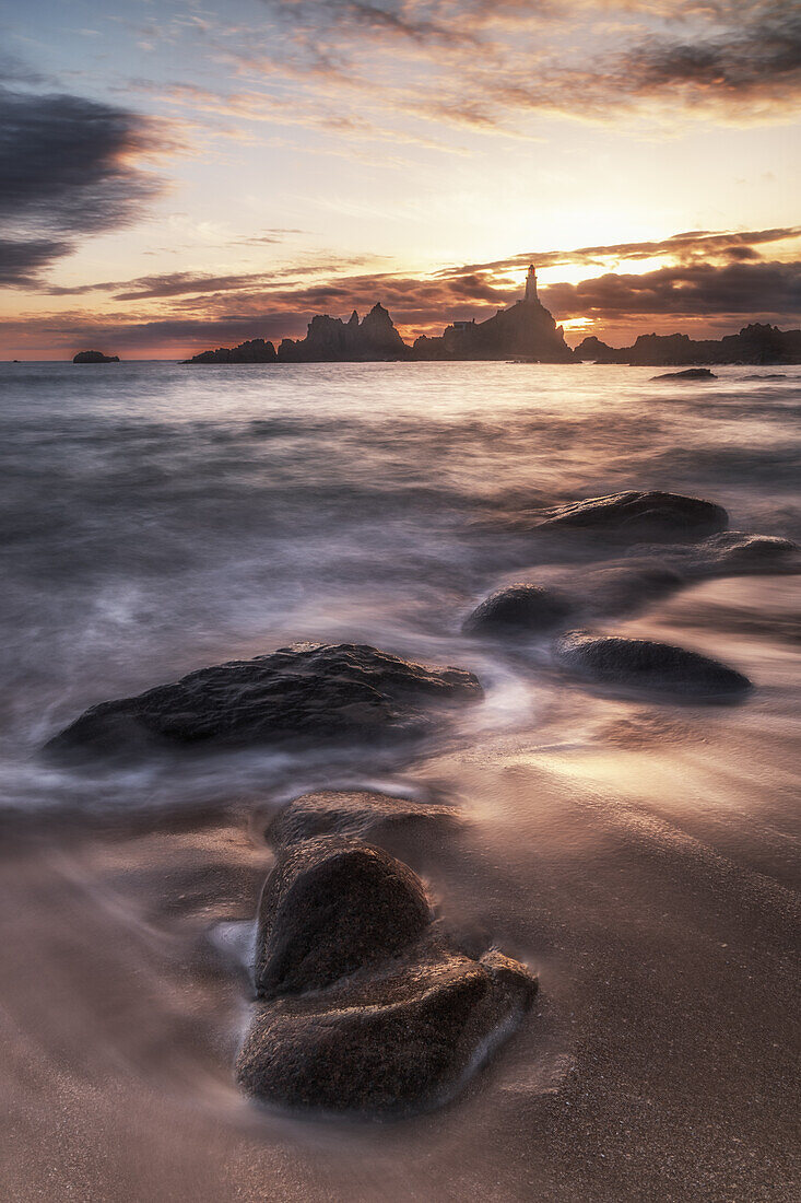  Le Corbiere lighthouse at sunset during low tide. Waves and stones on the beach. 