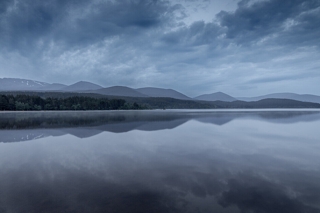  Mountain range reflected in Loch Morlich, Highlands, Scotland, UK. Overcast sky. Blue hour, morning. 