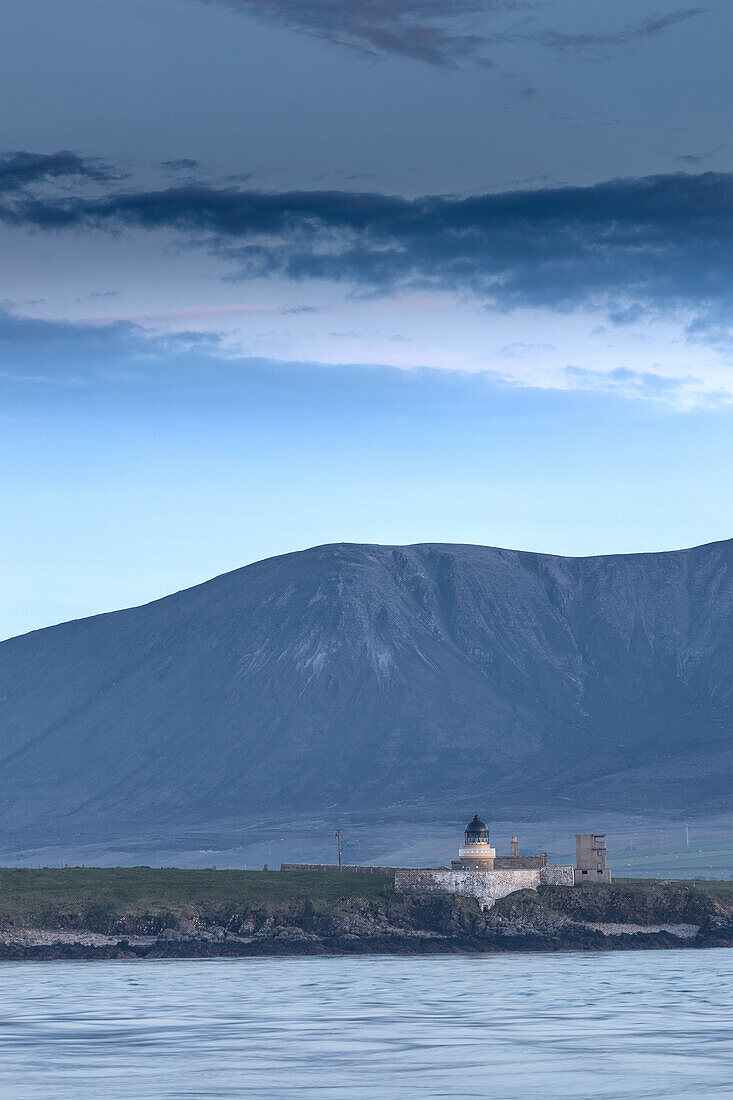  Graemsey low lighthouse in the evening light in front of mountains. Calm sea in the foreground. 