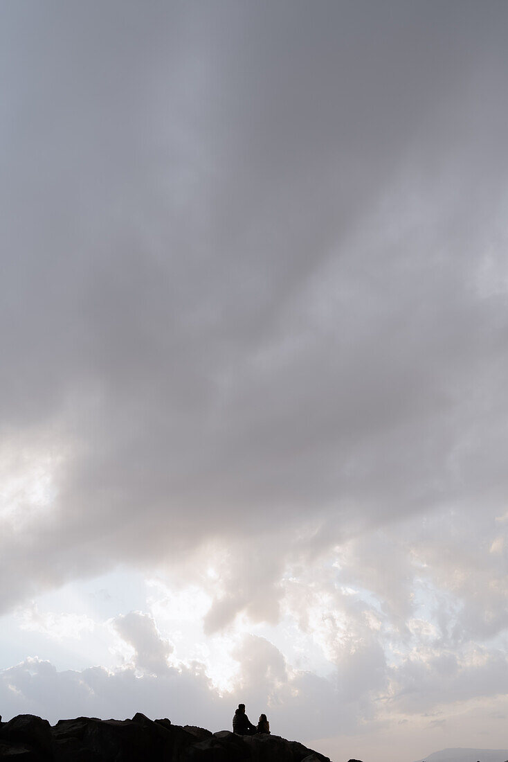 Scenic view of two lovers meeting on rocks by the sea, under a vast and moody sky.