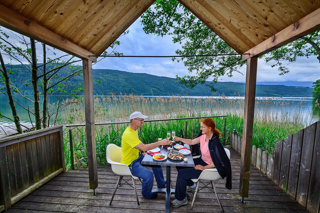  Man and woman sitting on the terrace of a tiny house and toasting each other, reed bivouac, Via paradiso, Millstätter See, Nockberge, Niedere Tauern, Carinthia, Austria 