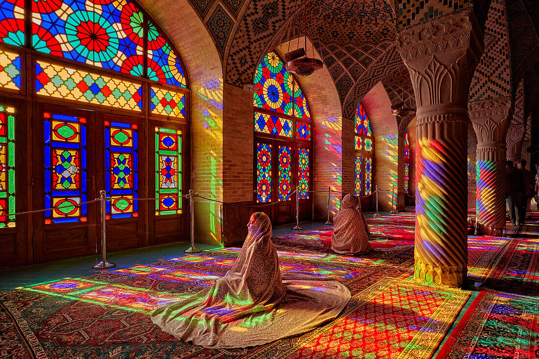 A woman sits in the light from multi-colored stained-glass windows inside the 19th century Nasir al-Mulk Mosque, aka the Pink Mosque. Shiraz, Iran.