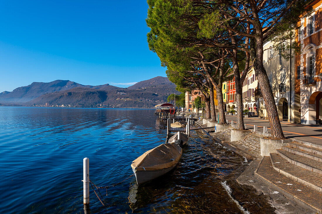 Altstadt mit Gebäuden und Bäumen am Ufer, Luganersee mit Boot an einem sonnigen Tag mit Bergen in Morcote, Tessin, Schweiz.