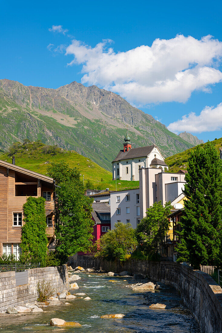 Altstadt mit Häusern und einer Kirche und dem Fluss Reuss und mit Bergen an einem sonnigen Sommertag in Andermatt, Uri, Schweiz.