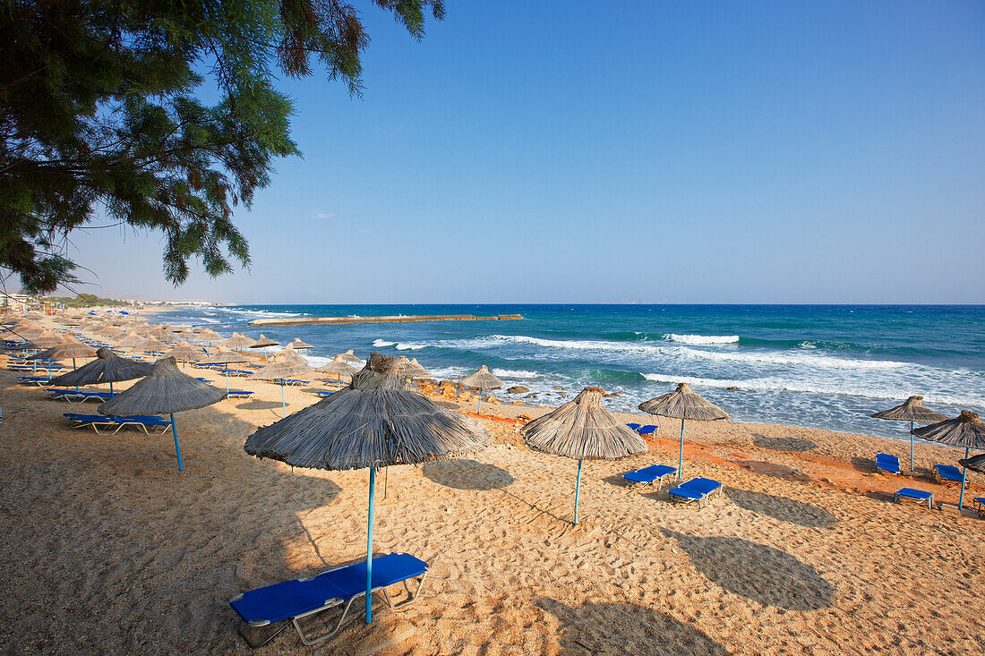  Liegestühle unter Strohsonnenschirmen an einem Sandstrand in der Nähe von Hersonissos, Kreta, Griechenland. 