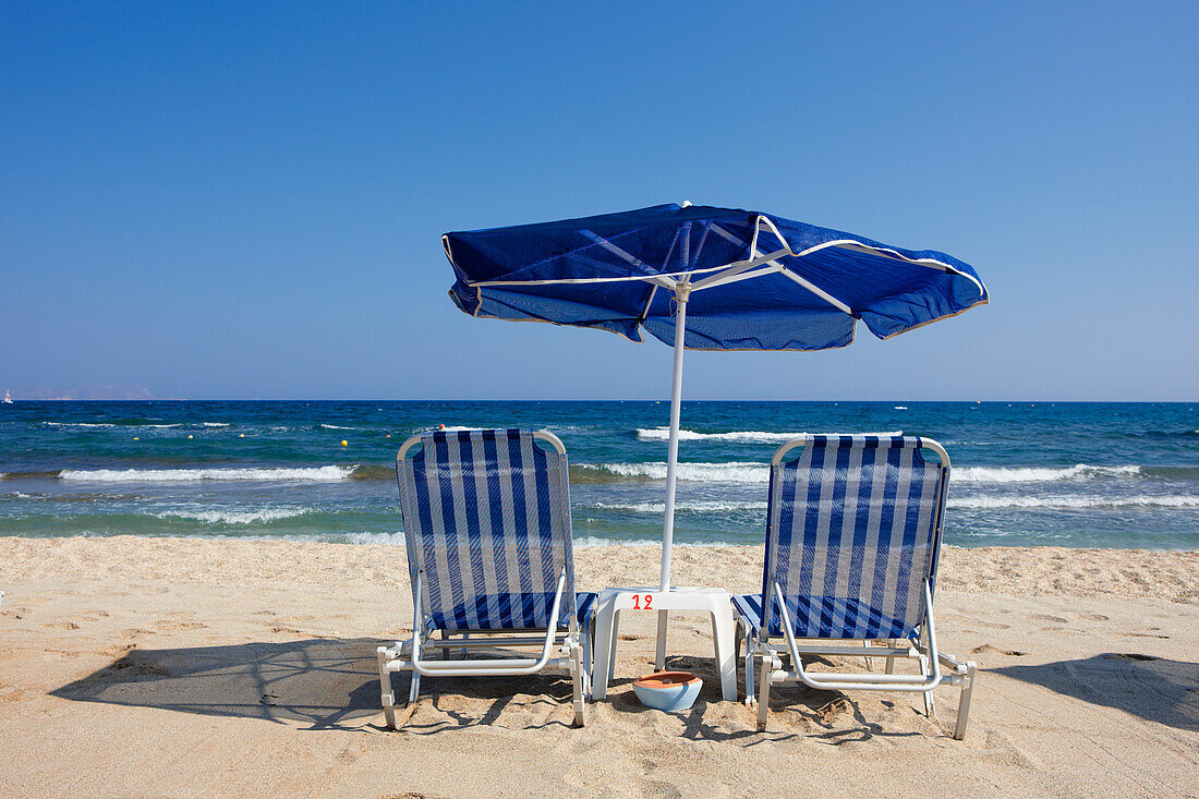  Zwei Liegestühle unter einem blau-weißen Sonnenschirm an einem Sandstrand in der Nähe von Hersonissos, Kreta, Griechenland. 