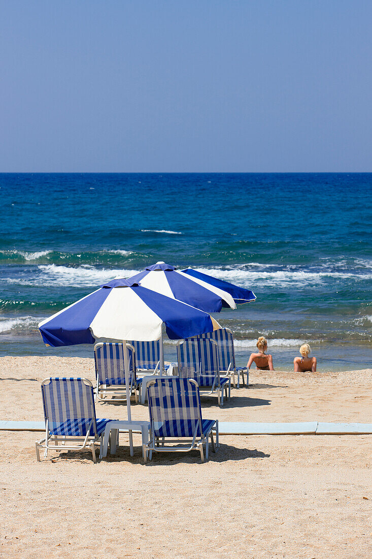 Loungers under blue and white parasols on a sandy beach near Hersonissos, Crete, Greece.