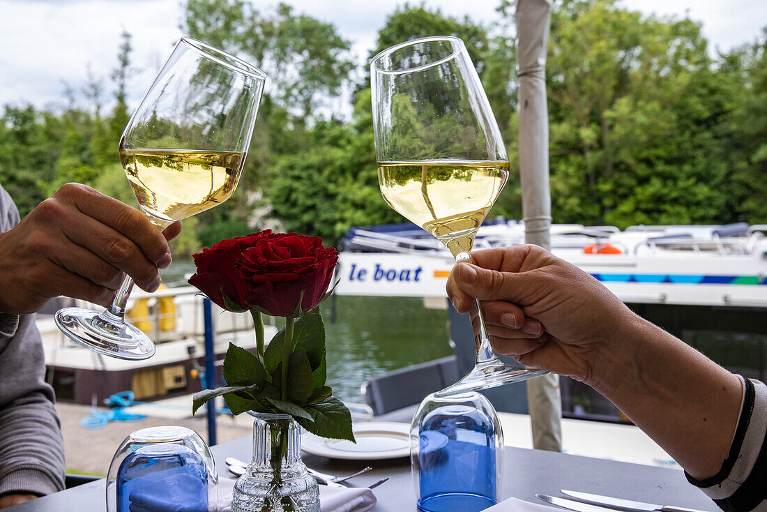  Hands of people toasting with white wine glasses in the beautiful Mediterranevm restaurant on the River Thames with Le Boat Horizon 4 houseboat moored behind, Bray, Berkshire, England, United Kingdom, Europe. 