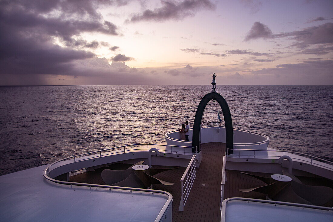 Paar im Krähennest auf dem Bug des Expeditionskreuzfahrtschiffs SH Diana (Swan Hellenic) mit Aldabra am Horizont in der Abenddämmerung, auf hoher See, in der Nähe der Seychellen, Indischer Ozean